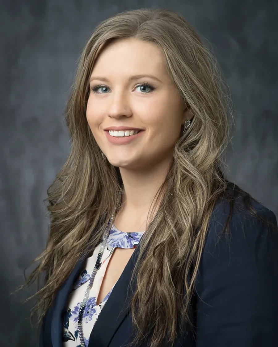 Studio headshot of a woman in a navy blazer against a mottled grey backdrop