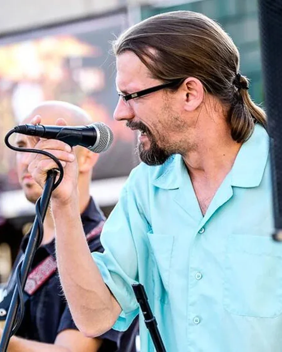 A singer in a pale turquoise shirt leans into a handheld microphone mid-song, a bandmate behind him