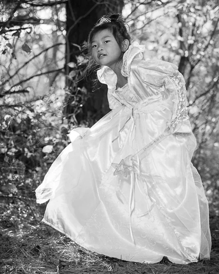 Black and white portrait of a young girl in a satin ball gown and tiara, holding a star wand beside a bare tree