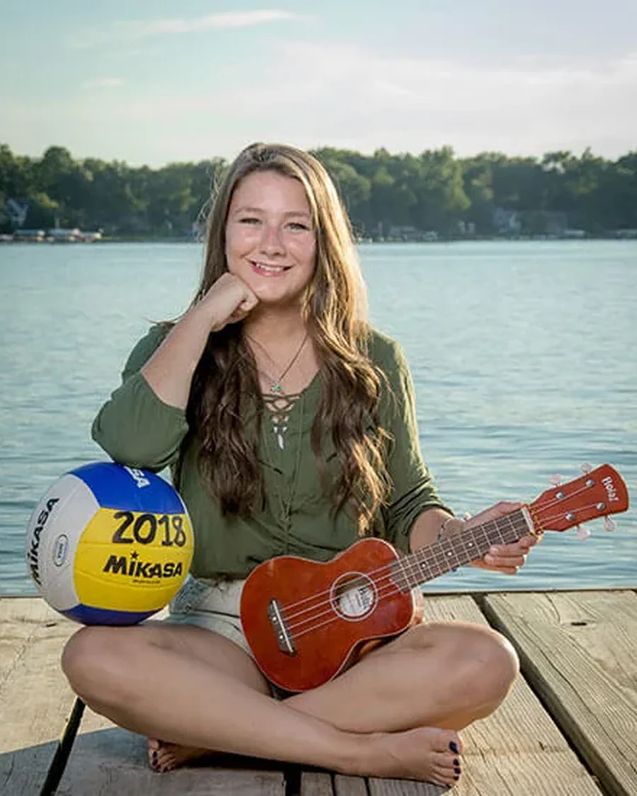 A high school senior sits cross-legged on a lake dock at golden hour, a volleyball at her side and a ukulele on her lap