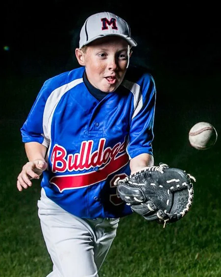 A youth baseball player in a blue Bulldogs jersey reaches out to catch the ball in his mitt under floodlights