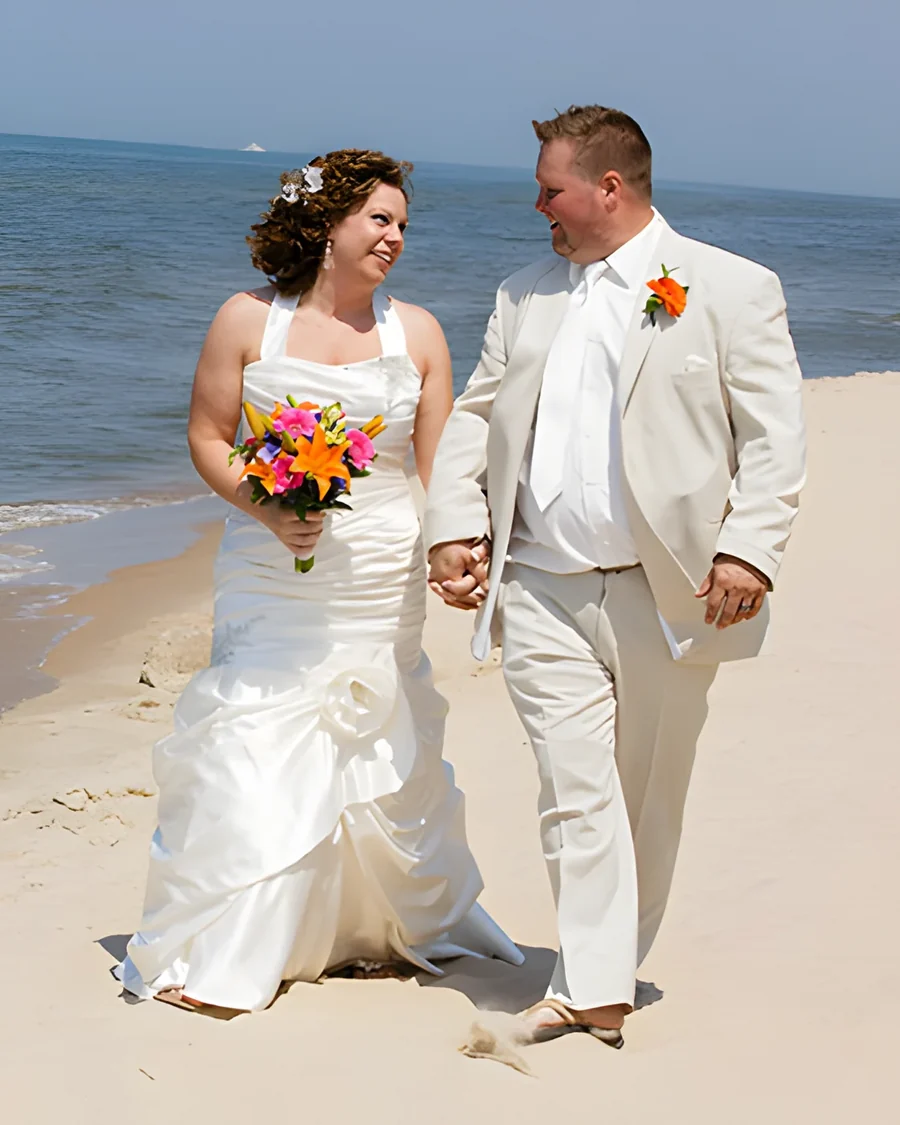 A bride and groom in ivory walk hand in hand along a sandy shoreline, her bouquet in bright tropical colours