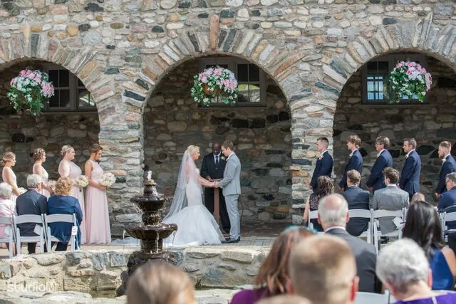 A wedding ceremony in front of a stone archway hung with flower baskets, the couple holding hands at the altar while guests watch from either side