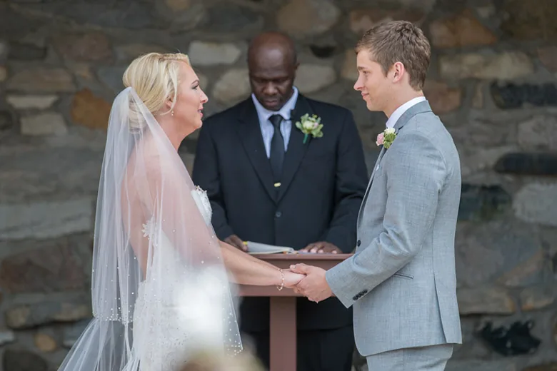 A bride in a long veil and a groom in a grey suit holding hands and smiling at each other during their ceremony, the officiant reading between them