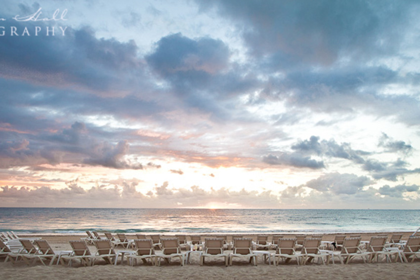 Rows of white loungers set out on a beach at sunrise, facing a calm sea under a wide pink and grey cloudy sky