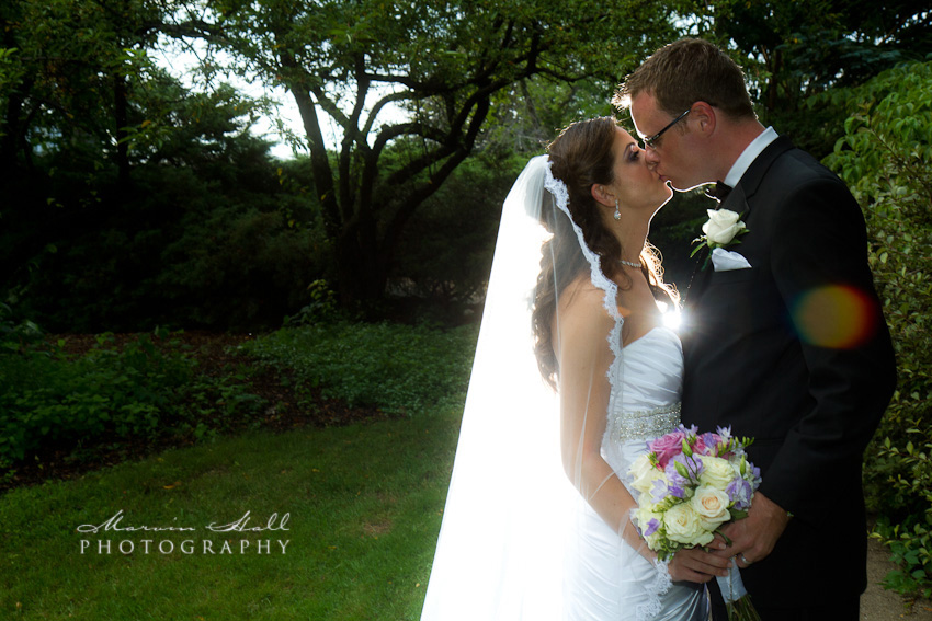 A bride and groom kissing in a green garden, her long veil lit from behind by the low sun, a pastel bouquet in her hand